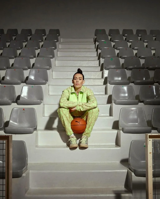 Adult female sits on arena stairs in patterned tracksuit holding basketball. Gray stadium seats receding into distance under even lighting.