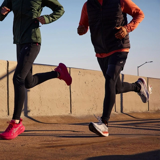 Two people in activewear running on an outdoor path. They wear colorful athletic shoes, with sky and a concrete barrier visible.