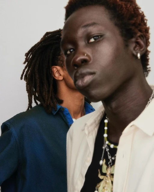 Two dark-skinned men in a fashion portrait, one with dreadlocks and a blue shirt, the other with light-colored shirt and necklace.