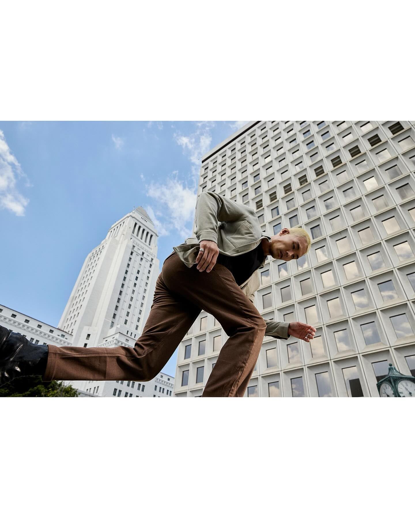 Low angle of an adult male running in brown pants and a gray shirt with buildings in the background during the daytime with cloudy sky.