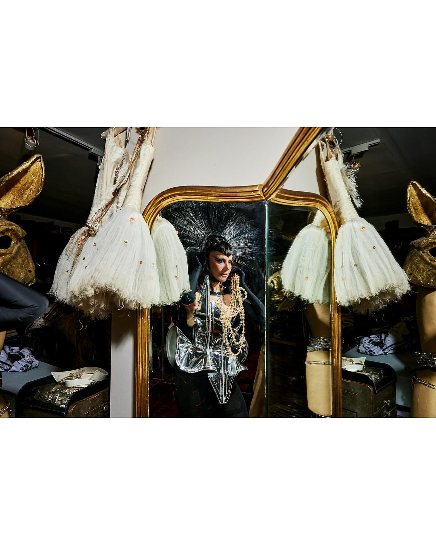 Woman in futuristic silver and black costume with pearl necklace, standing in cluttered interior next to a gold-framed mirror and dresses.