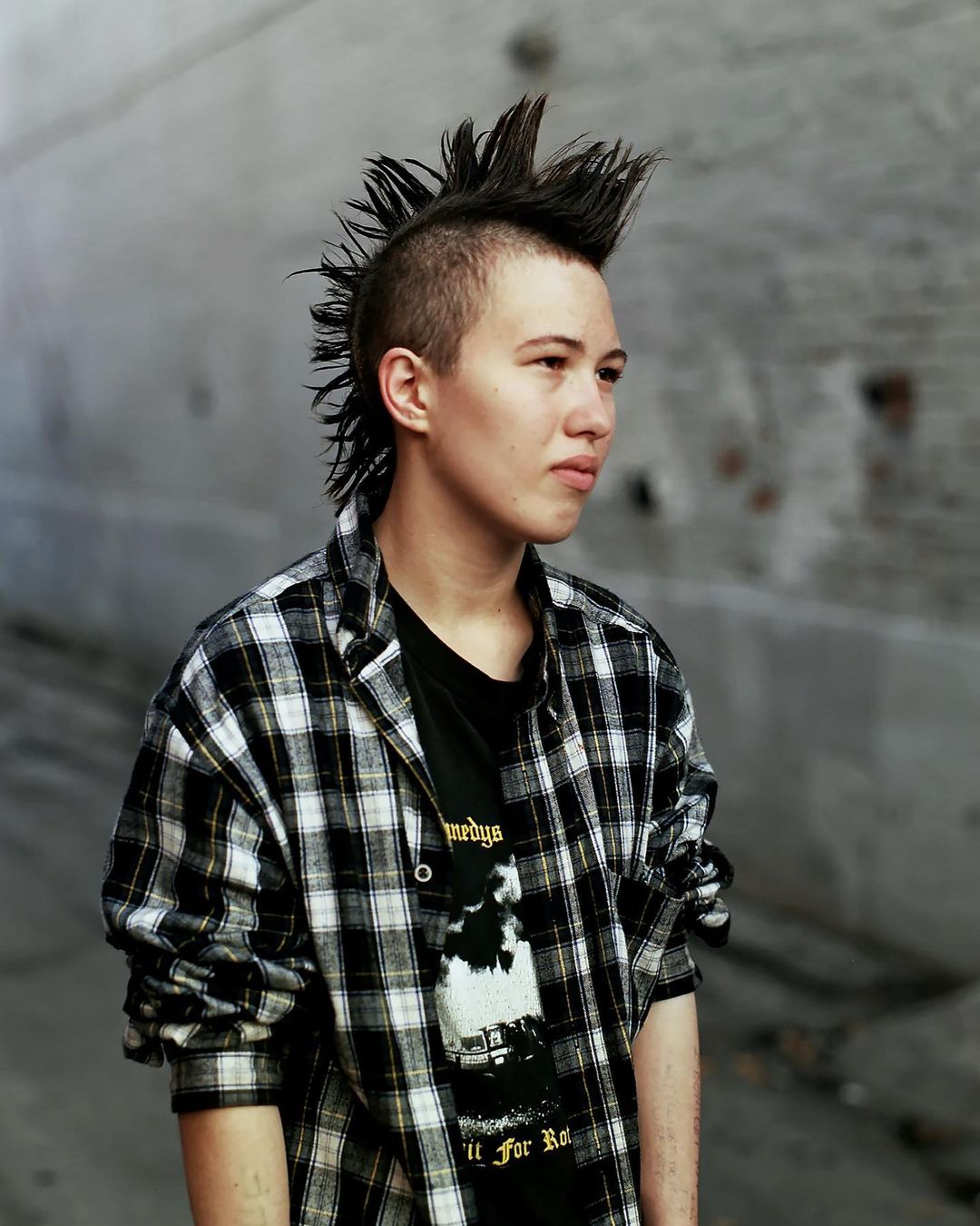 Portrait of a young adult with a mohawk hairstyle, wearing a black t-shirt and a plaid shirt, standing against a grey wall.