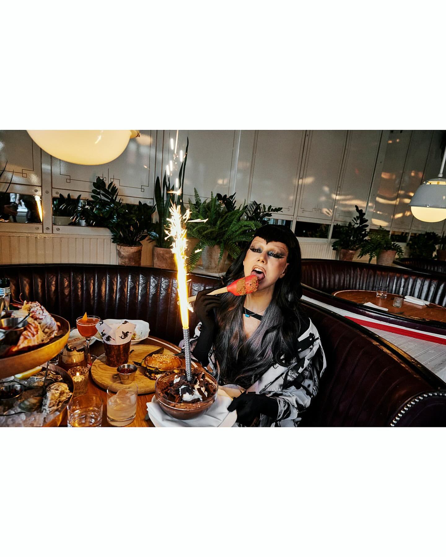 Person eating dessert with sparkler at indoor restaurant with mood lighting and dark wood booth seating, food and beverages on table.