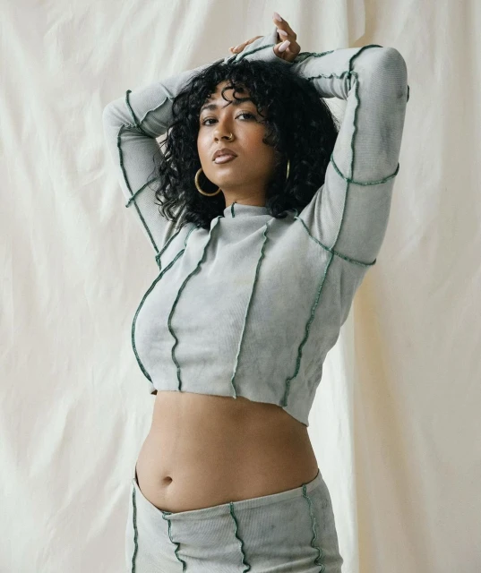 Portrait of a young adult woman with curly dark hair wearing a matching light green crop top and skirt with dark green stitching, indoors.
