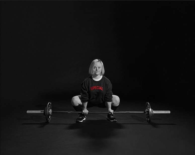 Woman with Down syndrome performing a barbell squat in a studio setting, wearing a black sweatshirt with the word 'SPECIAL' in red text.