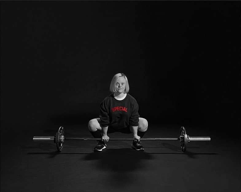 Woman with Down syndrome performing a barbell squat in a studio setting, wearing a black sweatshirt with the word 'SPECIAL' in red text.