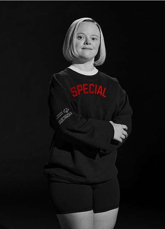 Portrait of an adult woman with Down syndrome in a black long-sleeved shirt and shorts, standing in front of a dark background.