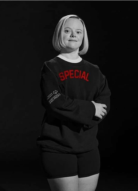 Portrait of an adult woman with Down syndrome in a black long-sleeved shirt and shorts, standing in front of a dark background.