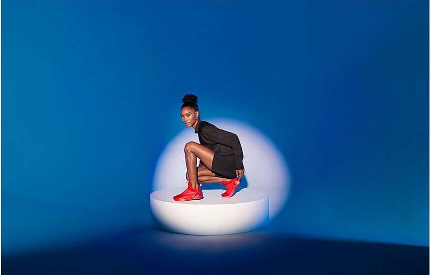 Elegant young woman with a slender build poses in an expressive squat on a white platform wearing an all-black outfit and red sneakers.
