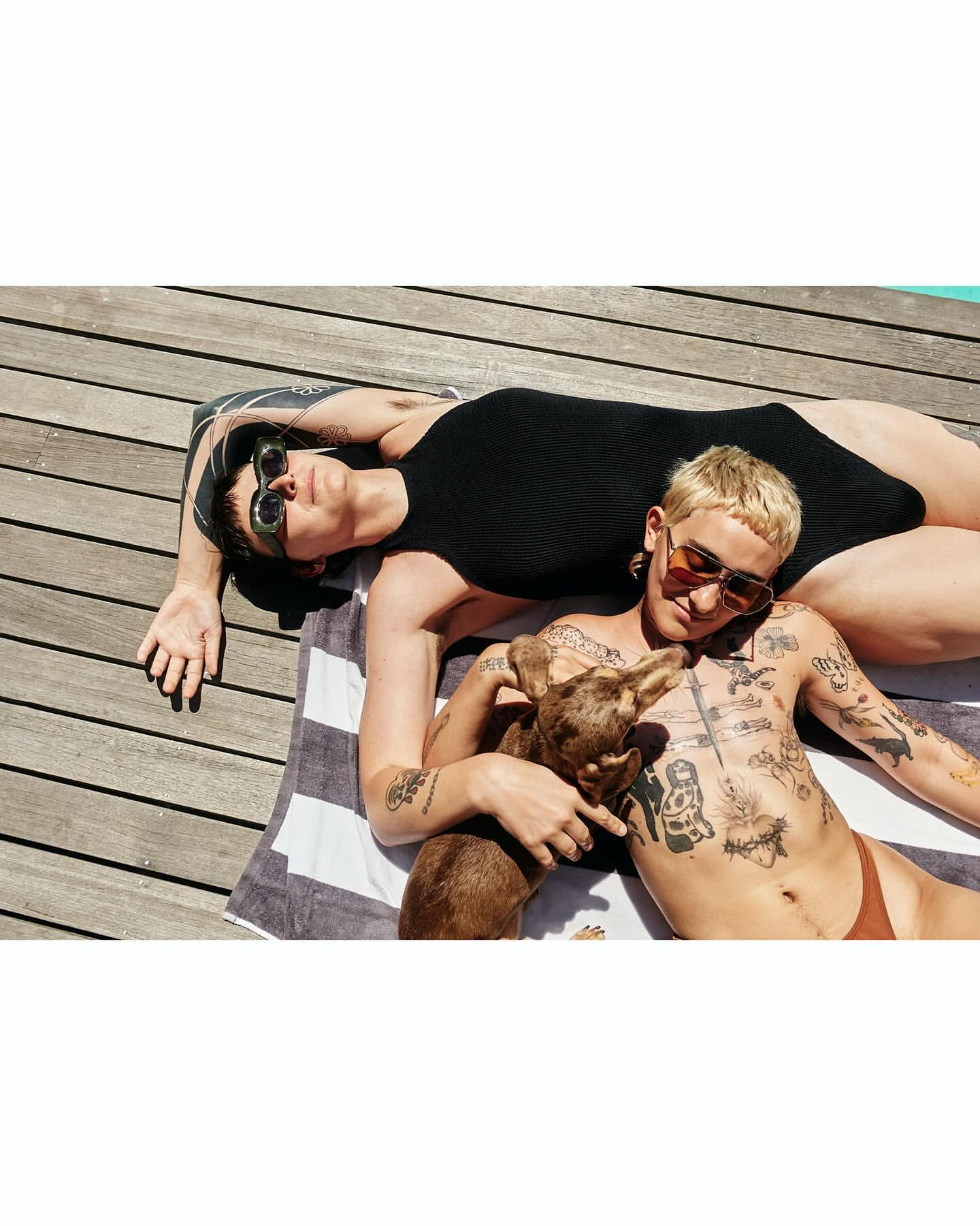 Two adults sunbathing on a towel with a dog on a wooden deck, wearing sunglasses and swimwear in bright sunlight, outdoor setting.