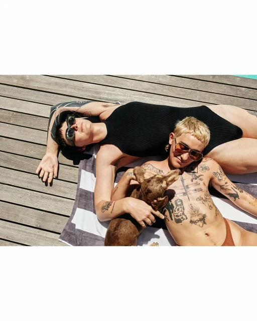 Two adults sunbathing on a towel with a dog on a wooden deck, wearing sunglasses and swimwear in bright sunlight, outdoor setting.