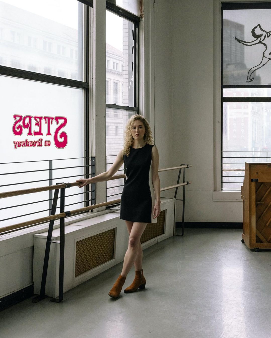 Young woman in black dress and brown ankle boots poses in dance studio with ballet barre and large window with dance studio signage.