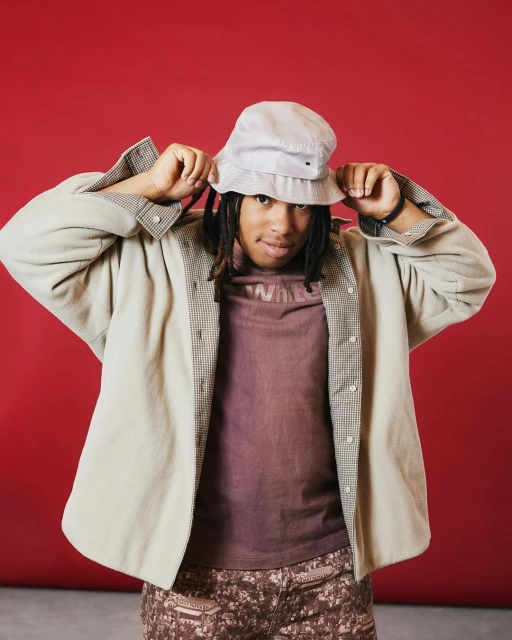Stylish young adult with dreadlocks and a bucket hat poses in front of a vibrant red backdrop, wearing a patterned shirt and jacket.