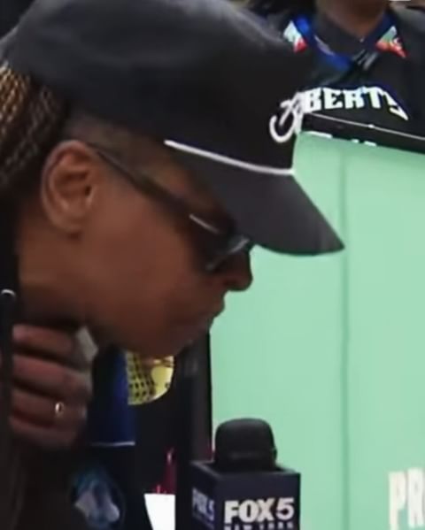 Close-up of an adult woman wearing a black baseball cap and sunglasses, speaking into a microphone at a press conference.