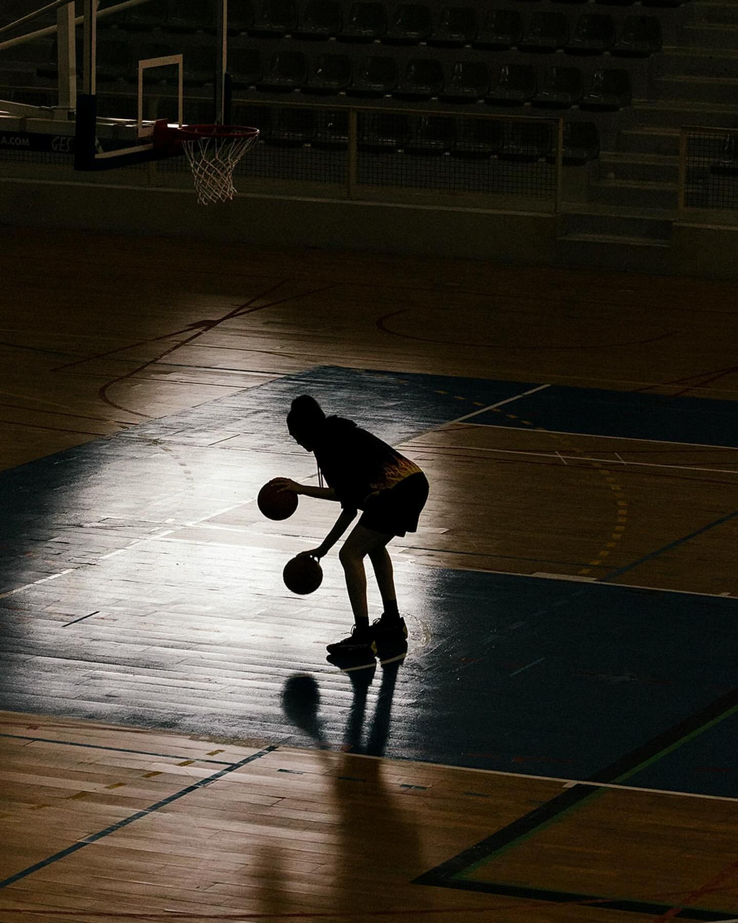 A basketball player is dribbling two basketballs in a gymnasium. A hoop and the empty stadium seating is visible in the background.