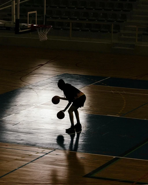 A basketball player is dribbling two basketballs in a gymnasium. A hoop and the empty stadium seating is visible in the background.