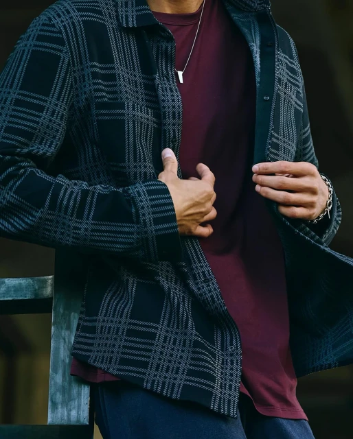 Man in a maroon tee and plaid shirt unbuttons the top button. A silver necklace adorns his neck in the soft, natural light.