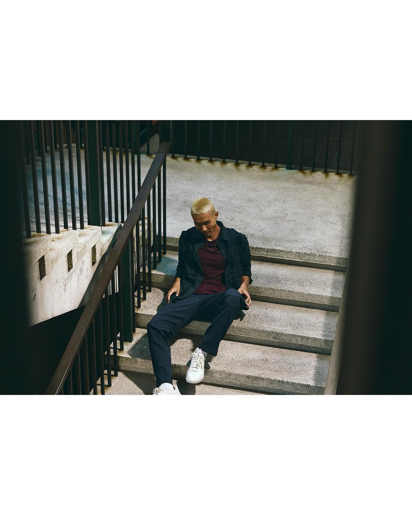 A young adult male with short blond hair sits on concrete steps outdoors, wearing a burgundy T-shirt, dark jacket, navy pants, and white sneakers.