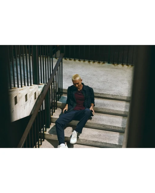 A young adult male with short blond hair sits on concrete steps outdoors, wearing a burgundy T-shirt, dark jacket, navy pants, and white sneakers.