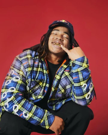 Young adult male in cap and plaid jacket crouches against red background showing gold teeth; studio shot for music or youth culture.
