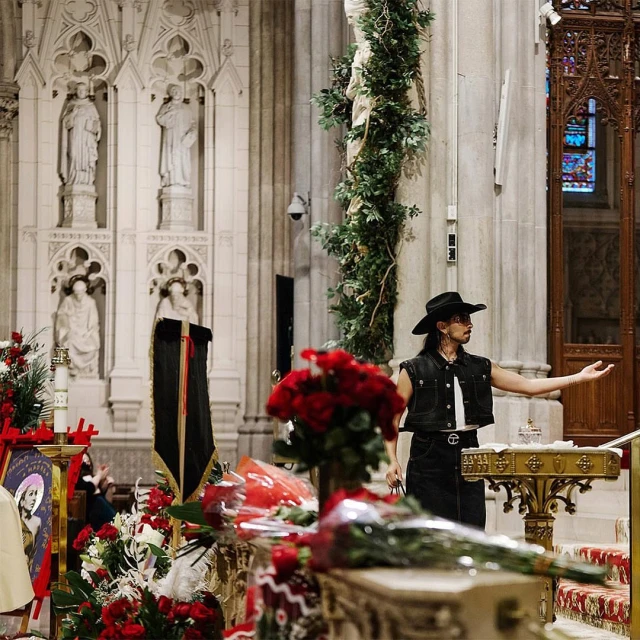 Man in vest and hat gesturing in church with red roses, black banner, statues, and stained glass windows in background.