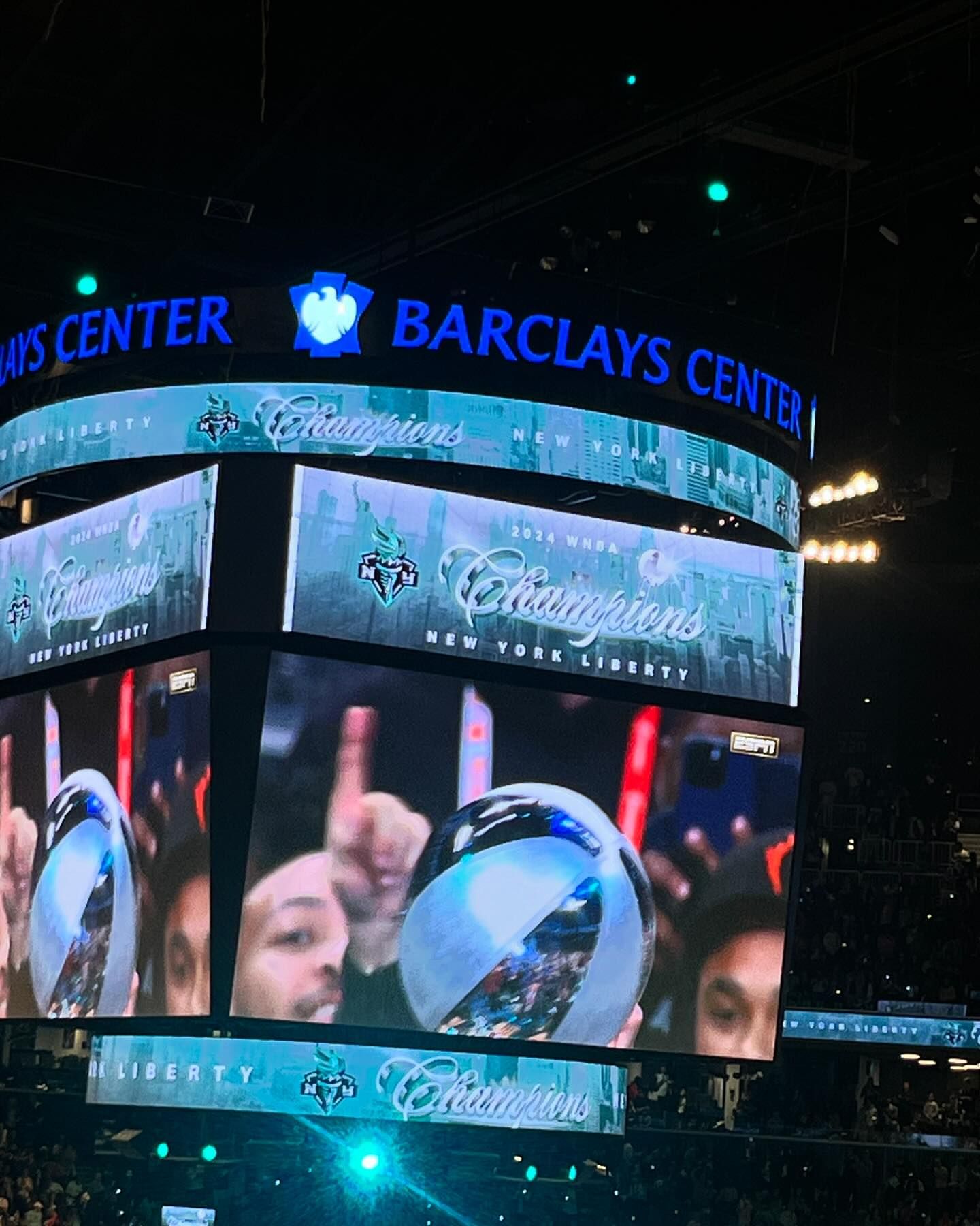 Interior view of Barclays Center screens displaying New York Liberty 'Champions' over a crowd, emphasizing sports and celebration.