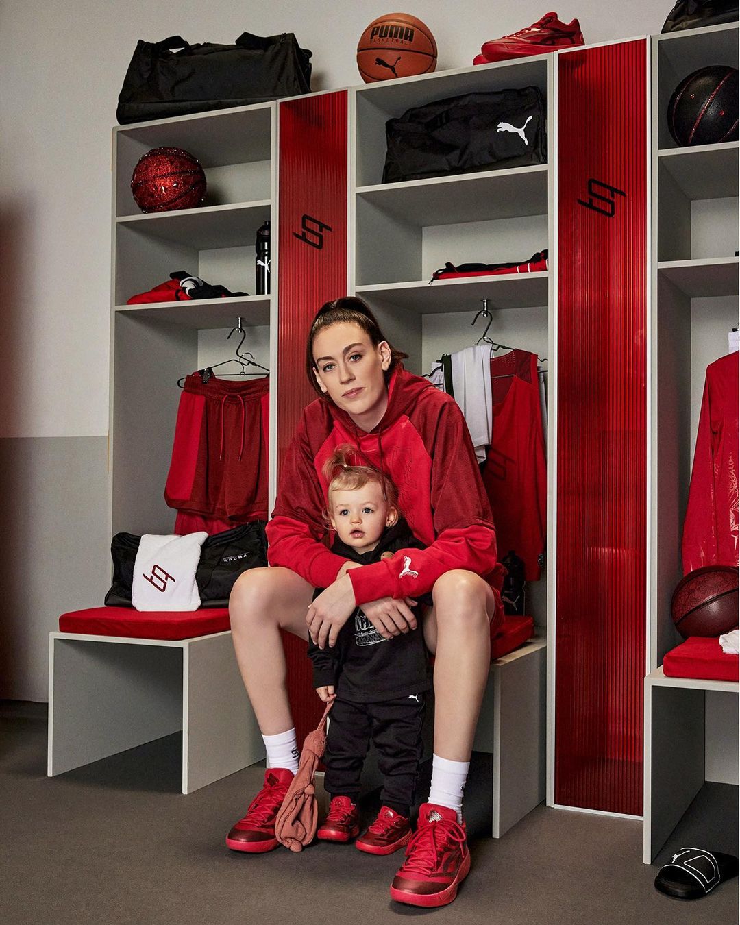 Adult female and child in matching red sneakers sit on a bench in a locker room, surrounded by sports equipment and clothing.