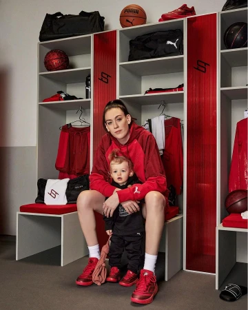 Adult female and child in matching red sneakers sit on a bench in a locker room, surrounded by sports equipment and clothing.