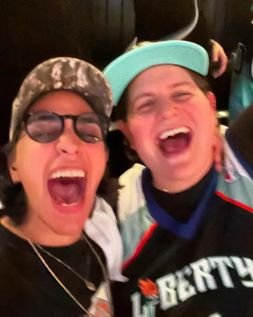 Two young adults joyfully shout and laugh while wearing casual attire and ballcaps at an indoor event with a dark background.
