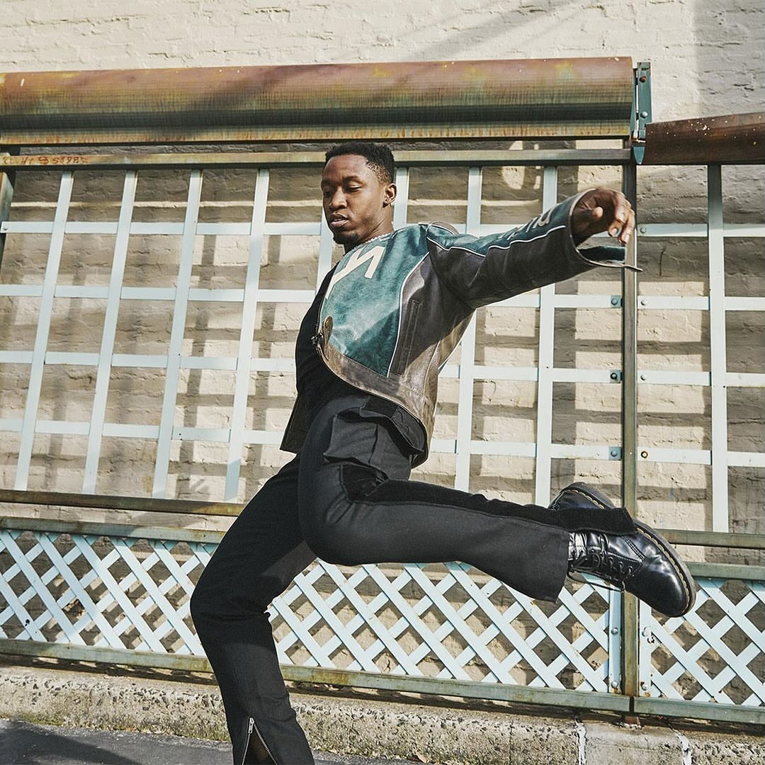 Stylish man mid-air wearing a vintage leather jacket, black pants, and boots, captured in an urban outdoor setting, dynamic pose.