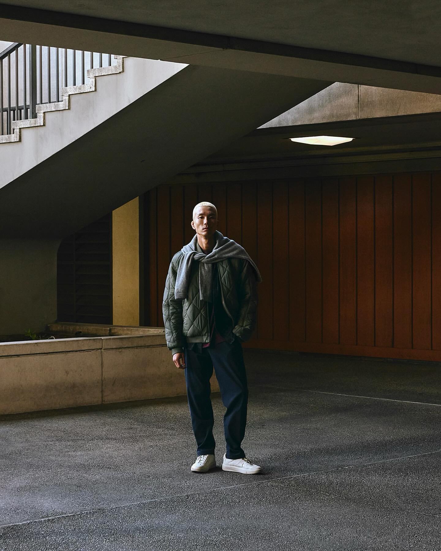 Full-length shot of an adult male standing in a covered outdoor area, wearing a quilted jacket, scarf, and sneakers.