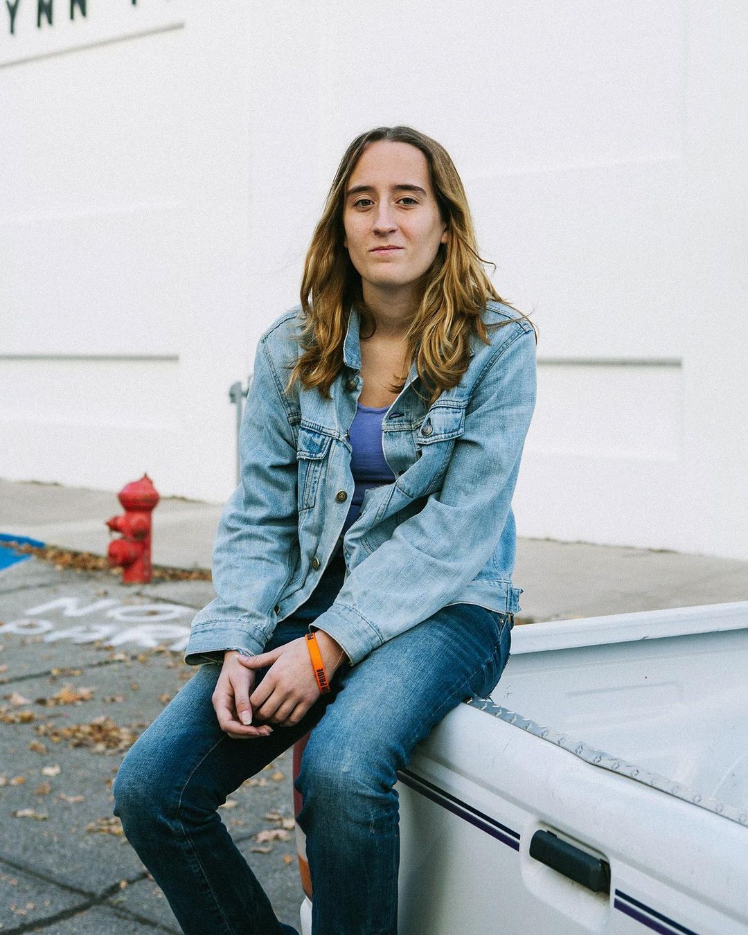 An adult female with shoulder-length hair sits on a white vehicle, wearing a denim jacket and jeans, captured in an outdoor setting.