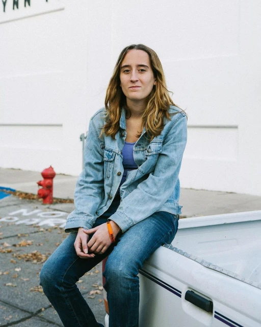 An adult female with shoulder-length hair sits on a white vehicle, wearing a denim jacket and jeans, captured in an outdoor setting.