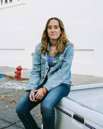 An adult female with shoulder-length hair sits on a white vehicle, wearing a denim jacket and jeans, captured in an outdoor setting.