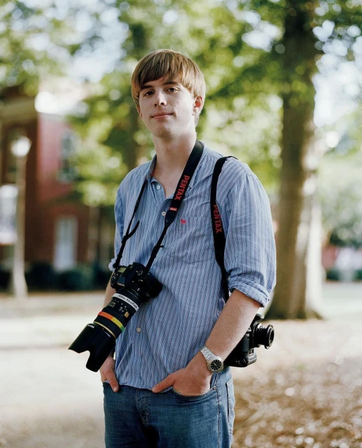A young adult male photographer with a DSLR camera and Pentax strap stands outdoors in casual clothing against a blurred background.
