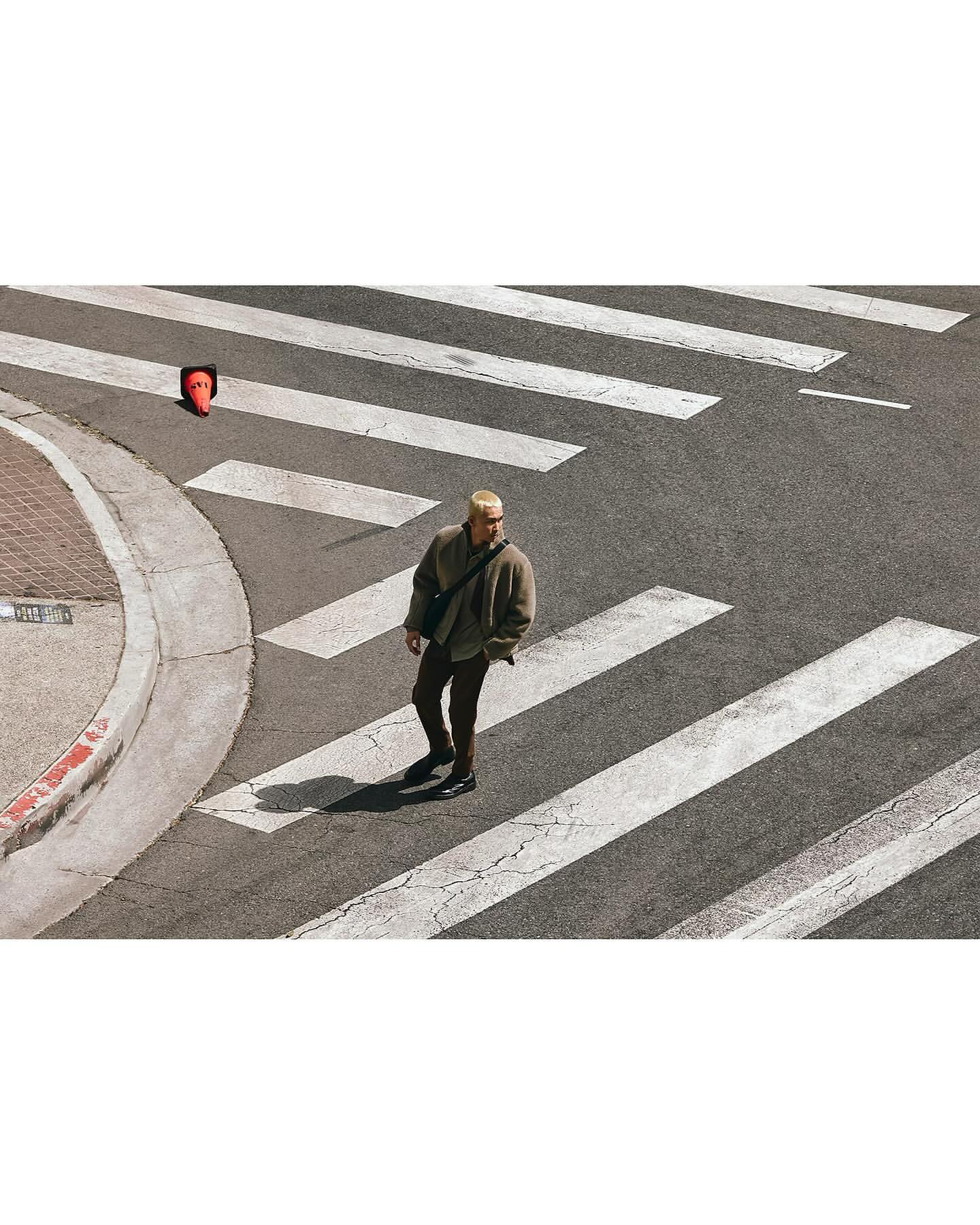 Adult male walks across a crosswalk with an orange traffic cone on the sidewalk, captured with an eye-level, wide shot, outdoor setting.