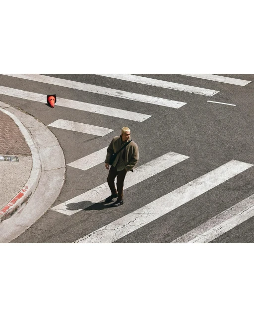 Adult male walks across a crosswalk with an orange traffic cone on the sidewalk, captured with an eye-level, wide shot, outdoor setting.