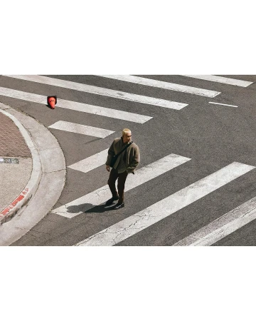 Adult male walks across a crosswalk with an orange traffic cone on the sidewalk, captured with an eye-level, wide shot, outdoor setting.