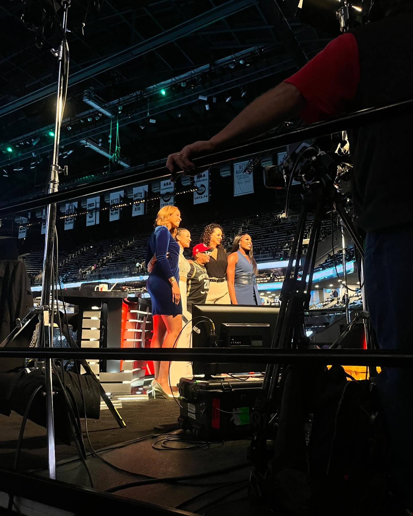 Group of diverse women on a sports broadcast set, medium shot with broadcasting equipment and stadium seating in the background.