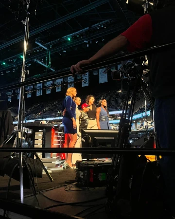 Group of diverse women on a sports broadcast set, medium shot with broadcasting equipment and stadium seating in the background.