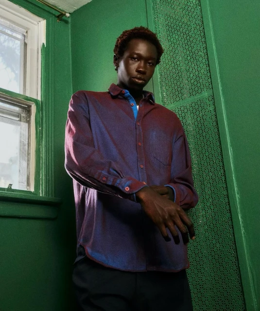 Adult male with dark skin stands indoors by a window, wearing a reddish-blue button-down shirt and dark pants in a portrait-style shot.