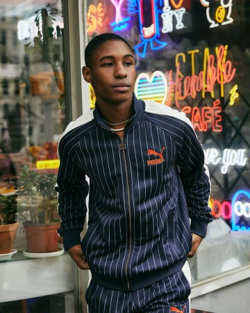 A young adult male poses in a navy and white striped Puma tracksuit against a backdrop of colorful neon cafe signs.