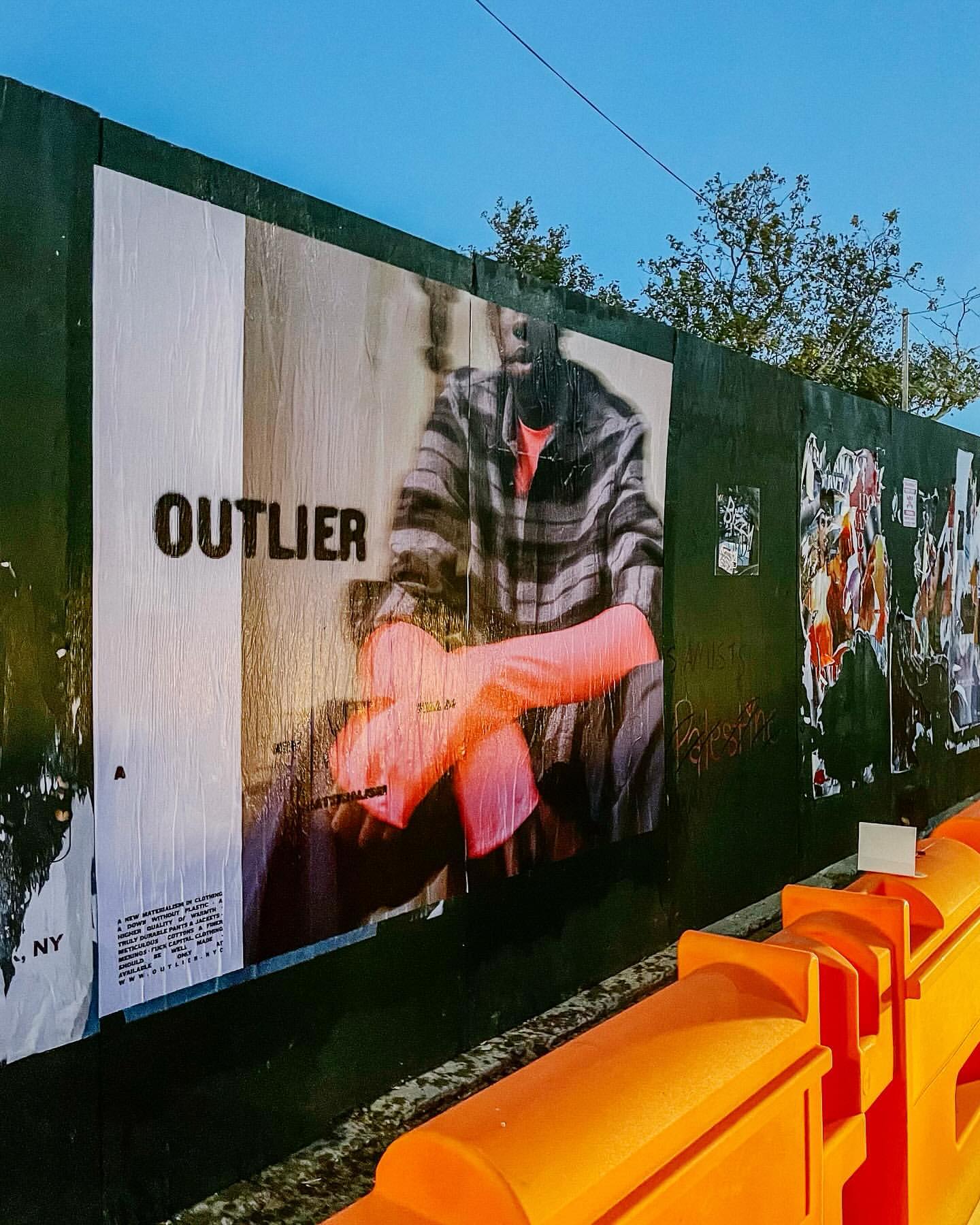Outdoor street art on a construction fence featuring a torn poster of a person in gloves and a striped shirt, with 'OUTLIER' printed on it.