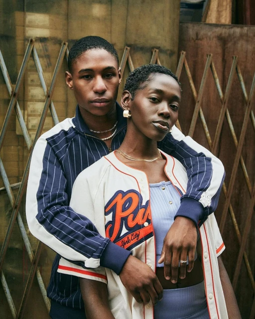 A stylish portrait of two young adults, one in a baseball jacket, embracing against a textured metal gate backdrop. Fashion and lifestyle theme.
