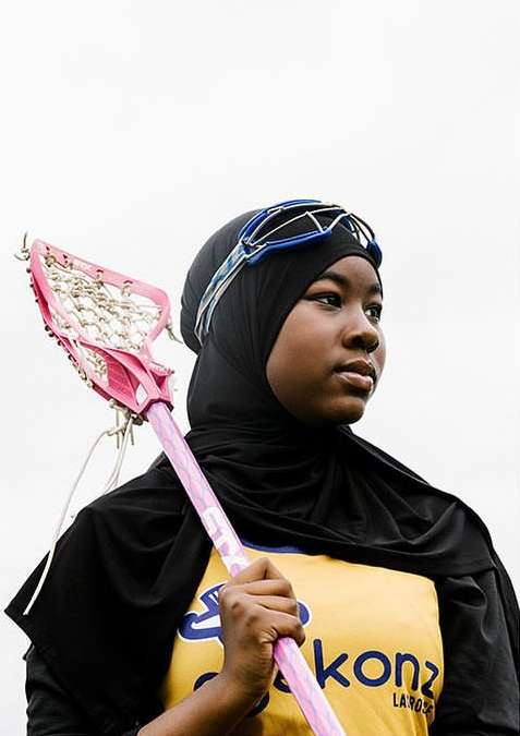 Young woman wearing a hijab and lacrosse goggles holds a pink lacrosse stick against a bright white sky backdrop in a medium portrait shot.