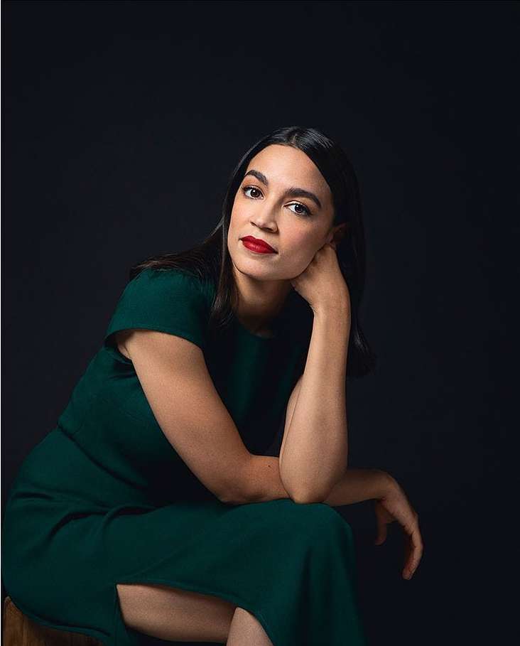 Elegant portrait of a woman with dark hair in a fitted dark green dress, seated against a dark background, showcasing a poised and confident pose.