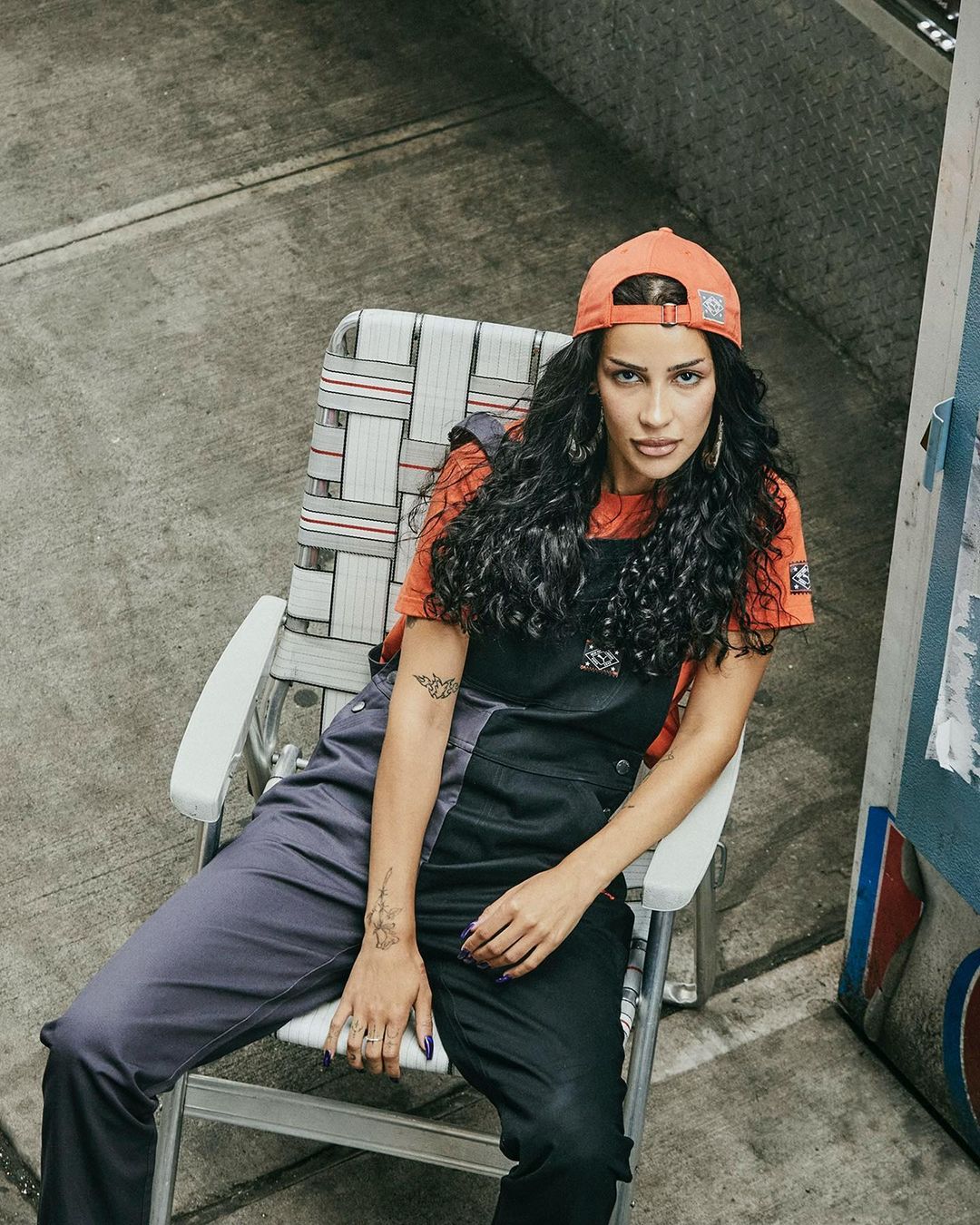 A woman with curly dark hair sits in a lawn chair wearing black overalls and an orange t-shirt, in an outdoor urban setting.