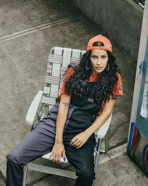 A woman with curly dark hair sits in a lawn chair wearing black overalls and an orange t-shirt, in an outdoor urban setting.