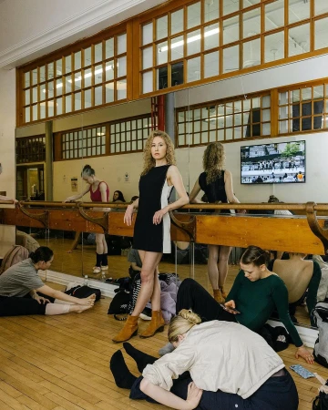 A woman in a black dress and tan ankle boots poses in a dance studio with other dancers, mirrors, and ballet barres.