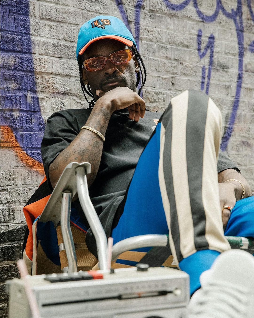 Black man wearing tinted orange sunglasses, blue cap, and striped athletic pants seated outdoors before a brick wall.